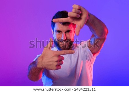 Cheerful caucasian guy making picture frame with fingers, capturing moment in photo and smiling to camera, standing over purple and blue neon studio backdrop. Professional photography concept