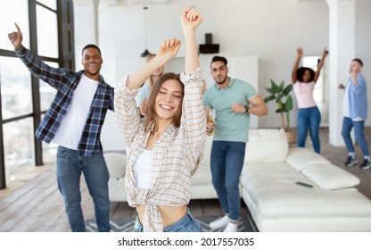 Cheerful Caucasian lady with her diverse friends dancing and drinking beer at home. Positive multiracial millennials moving to music and laughing, enjoying weekend together indoors - Powered by Shutterstock