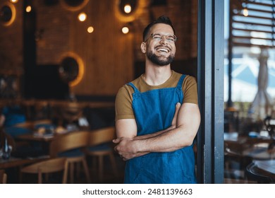 A cheerful Caucasian male bartender stands with arms crossed in a well-lit coffee house, dressed in a blue apron, indicative of his profession. - Powered by Shutterstock