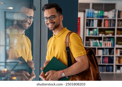 A cheerful Caucasian male student with glasses looks outward from a library window, a book in hand and a backpack slung over his shoulder, reflecting an engaging learning environment. - Powered by Shutterstock