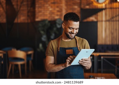 A cheerful Caucasian male waiter in an apron engages with a digital tablet inside a cozy restaurant, embodying efficient hospitality. - Powered by Shutterstock