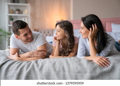 Cheerful caucasian man and his beautiful happy little daughter looking on each other while lying in bed with smiling mother