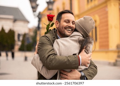 A cheerful Caucasian man and woman in winter clothing celebrate International Women's Day with hugs in a picturesque urban setting. The man holds a red rose. - Powered by Shutterstock