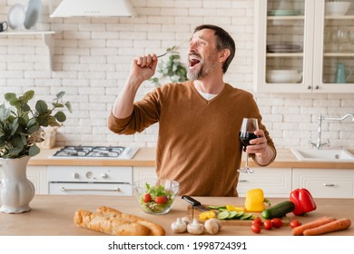 Cheerful caucasian mature middle-aged man father husband singing using spoon as a microphone while drinking red wine in the kitchen, preparing cooking vegetable salad - Powered by Shutterstock