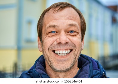 Cheerful Caucasian middle-aged man over 50, close-up smiling unshaven face with white teeth and blue eyes. A happy white male of fifty years old on a city street in Europe.