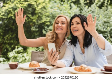 Cheerful Caucasian Middle-aged Mature Women Best Friends Greeting Waving Their New Friend While Sitting And Having Breakfast In City Cafe Using Smart Phone. Friendship Concept.