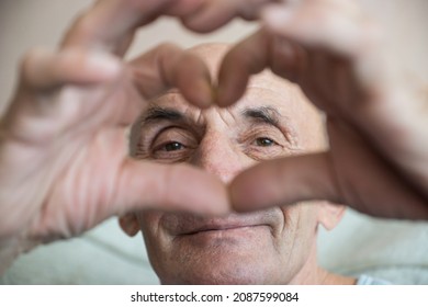 Cheerful Caucasian Senior Citizen 70 Years Old Portrait Smiling And Looking Through Hands Making Heart Gesture. Greetings With Love From An Older Person. Selective Focus, Shallow Depth Of Field