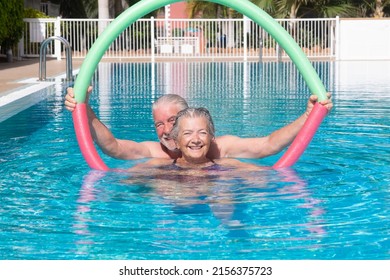Cheerful caucasian senior couple having fun in outdoors swimming pool doing exercise with swim noodles. Smiling retired people playing together in the pool water under the sun