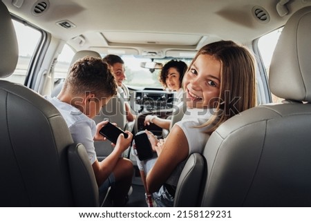 Cheerful Caucasian Teenage Girl Smiles Into Camera While Sitting in Minivan Car with Her Brother, Mother and Father, Happy Four Members Family Enjoying Weekend Road Trip