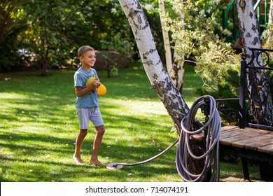 A Cheerful Caucasian Toddler Boy Having Fun Playing With Water Filled Balloons In Garden At Backyard.