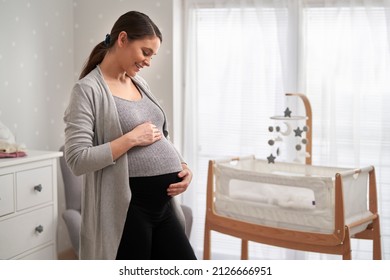 Cheerful Caucasian Woman In Advanced Pregnancy Standing In The Baby's Room And Stroking Her Abdomen