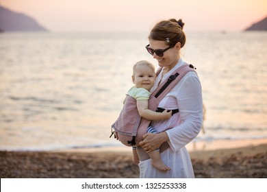 Cheerful Caucasian Woman With Baby Daughter In Buckle Carrier On Beach At Sunset