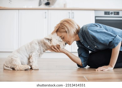 Cheerful caucasian woman leaning forward with her legs underneath body to cute dog's nose on kitchen background. Joyful young adult in denim shirt having fun with furry friend while training indoors. - Powered by Shutterstock