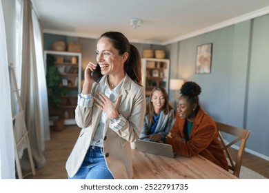 A cheerful Caucasian woman in professional attire engages in a phone conversation in her well-lit home office, with diverse colleagues in the background. - Powered by Shutterstock