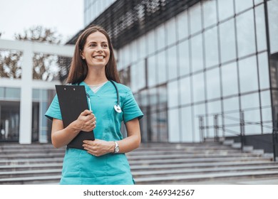 A cheerful Caucasian woman in scrubs holds a clipboard, displaying professionalism and readiness near a hospital setting. - Powered by Shutterstock