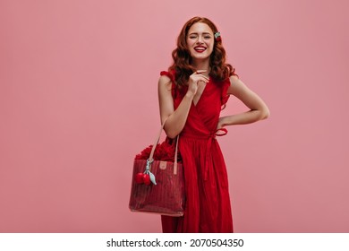 Cheerful Caucasian Young Girl Smiles Broadly Looking At Camera On Isolated Background. Wonderful Young Red-haired Lady In Red Long Dress With Bag Of Same Color. 