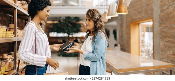 Cheerful ceramic store owner receiving an NFC payment from a customer in her shop. Female small business owner scanning a smartphone on a credit card machine while serving a customer. - Powered by Shutterstock