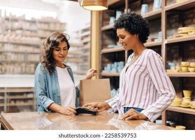 Cheerful ceramic store owner scanning a credit card on a card machine to receive a payment. Happy small business owner doing a cashless transaction while serving a customer at checkout.