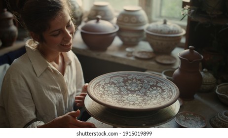 Cheerful ceramist playing with potters wheel in studio. Portrait of excited woman spending time in pottery. Closeup happy female artist examining plate with ornament in workshop. - Powered by Shutterstock