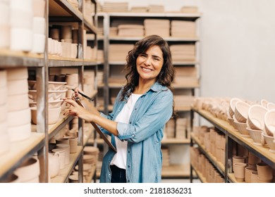 Cheerful ceramist smiling at the camera while stocktaking in her shop. Creative businesswoman managing a store with handmade ceramic products. Female entrepreneur running a successful small business.