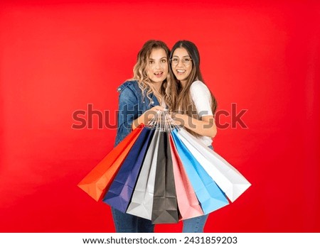 Cheerful charming attractive two best friends young women wear denim casual clothes holding paper package bags after shopping looking smiling camera isolated red background. Lifestyle concept.