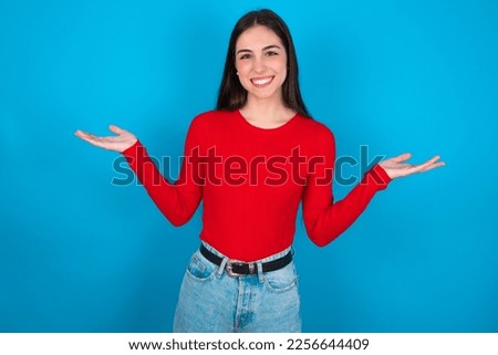 Cheerful cheery optimistic young caucasian brunette girl wearing red T-shirt against blue wall holding two palms copy space