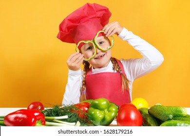 A cheerful child in a chef's cap has fun with vegetables and holds pepper in front of his eyes, as if in glasses. Little girl sits at a table on a background of yellow wall.