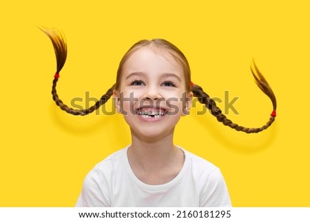 Cheerful child girl with growing teeth throws pigtails and laughs on a yellow background. Funny girl has fun and shows positive emotions by playing with her hair. Happy childhood concept