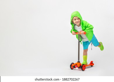 Cheerful Child Kid In Bright Green Jacket With Hood And Rubber Boots Rides Scooter On White Background With Copy Space. Studio Shot Toddler, Boy 4 Years Old. Childhood And Active Leisure Concept.