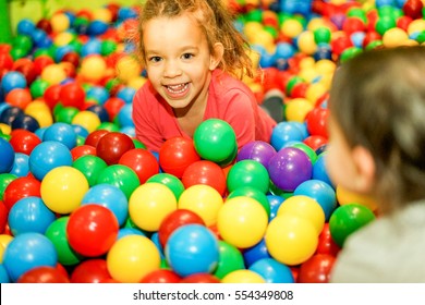 Cheerful children playing inside ball pit swimming pool - Happy people having fun in baby playground indoor - Childhood and friendship concept - Focus on left female face - Warm vivid filter