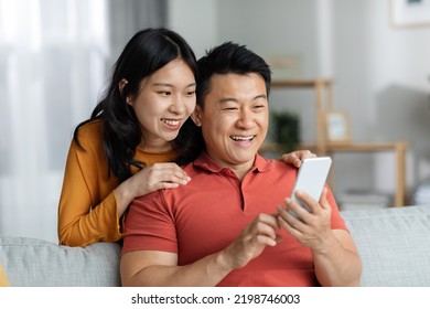 Cheerful Chinese Lovers Having Video Chat With Friends, Positive Asian Couple Making Online Party With Family While Staying Together At Home, Using Mobile Phone, Looking At Gadget Screen And Smiling