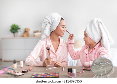 Cheerful chinese teenage girl and young female in pink pajamas and towels on heads do makeup in bedroom interior. Daughter applies cosmetics to mom face, lesson with cosmetics and bachelorette party
