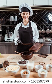 cheerful chocolatier in apron holding cake scraper near chocolate molds and bowl with melted dark chocolate