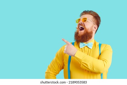 Cheerful Chubby Man Laughs Ridiculously Crazy Presenting New Promotion Or Offering Sale. Smiling Bearded Guy In Yellow Shirt And Suspenders Points His Finger At Copy Space On Light Blue Background.