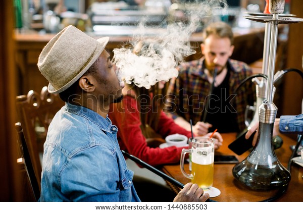 Cheerful company of friends, drinking beer and\
smoking hookah in shisha lounge. Rest in hookah, African man makes\
tornado out of smoke\
shisha