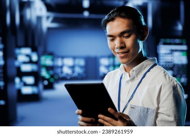 Cheerful computer operator in data center uses tablet to prevent system overload during peak traffic periods. BIPOC man in server facility ensuring enough network bandwidth for smooth operations