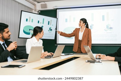 A cheerful and confident Asian businesswoman stands, present bar charts data from projector screen to her office colleagues. Asian business women leader role at the meeting. - Powered by Shutterstock