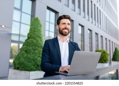 Cheerful Confident Handsome Young Caucasian Guy With Beard In Suit Typing On Laptop Near Modern Office Building. Successful Business Outdoor, Businessman And Conference Due Social Distance, New Normal