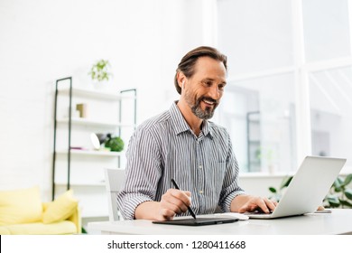 Cheerful Confident Middle Aged Man Sitting At The Table And Looking At The Screen Of His Laptop While Holding A Stylus In His Right Hand