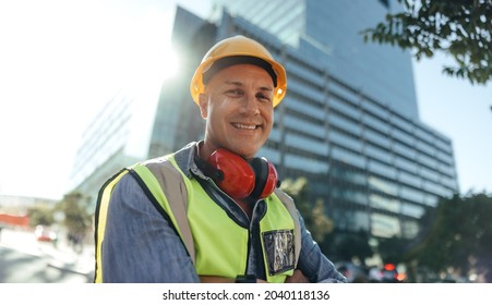 Cheerful construction worker smiling at the camera while standing with his arms crossed in the city. Mid-adult blue collar worker standing in front of high rise buildings in his workwear.