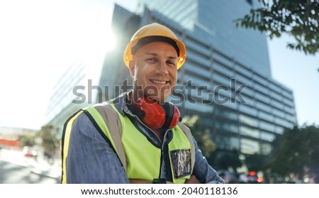 Cheerful construction worker smiling at the camera while standing with his arms crossed in the city. Mid-adult blue collar worker standing in front of high rise buildings in his workwear.