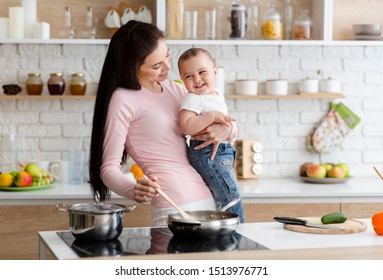 Cheerful Cooking. Millennial Woman Laughing With Baby Son, Preparing Food On Cooktop At Kitchen, Free Space