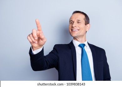 Cheerful, corporate, executive man in formal wear with tie having invisible screen, trying to touch a button with forefinger in front of him, standing over gray background
