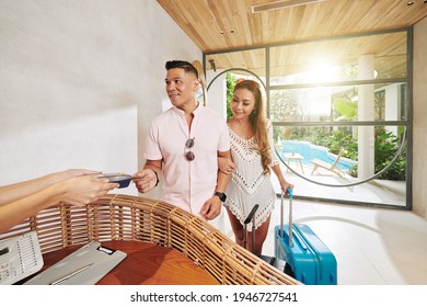 Cheerful Couple With Baggage Standing At Reception And Paying For Room In Spa Hotel