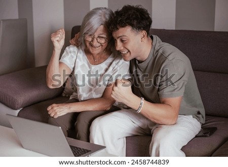 Cheerful couple of boy and elderly grandmother watching summer olympic games together on laptop rejoicing for victory sitting on comfortable sofa in living room