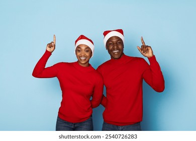 A cheerful couple dressed in matching red sweaters and Santa hats poses with raised hands, smiling brightly against a light blue background, embodying the spirit of the holiday season. - Powered by Shutterstock