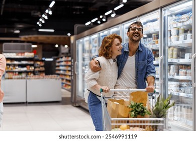 Cheerful couple embracing and laughing while choosing frozen food from refrigerated shelves, pushing shopping cart with fresh produce in supermarket - Powered by Shutterstock