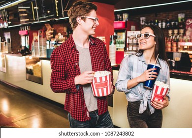 Cheerful couple in glasses are looking to each other and smiling. They are posing. Girl has a cup of coke and a basket of popcorn while guy is holding only basket of popcorn. - Powered by Shutterstock