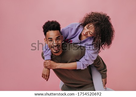Cheerful couple having fun on pink background. Girl sitting on her boyfriends back. Pretty dark-skinned woman hugging man dressed in brown shirt