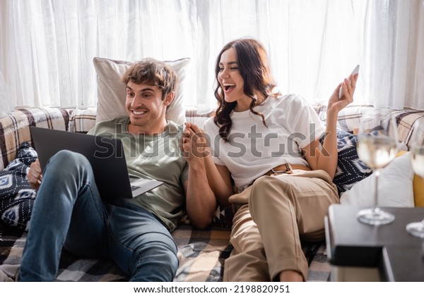 Cheerful couple holding hands while using\
devices near blurred wine in camper\
van